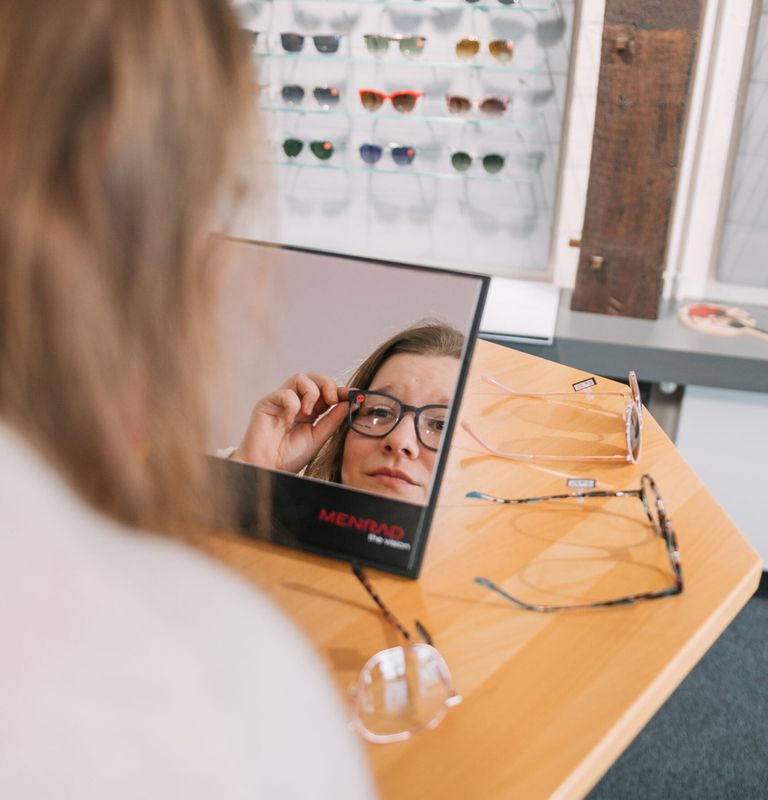 Eine Person betrachtet eine Sonnenbrille im Spiegel. An der Wand dahinter sind verschiedene Modelle ausgestellt und mehrere Modelle liegen auf einem Holztisch.