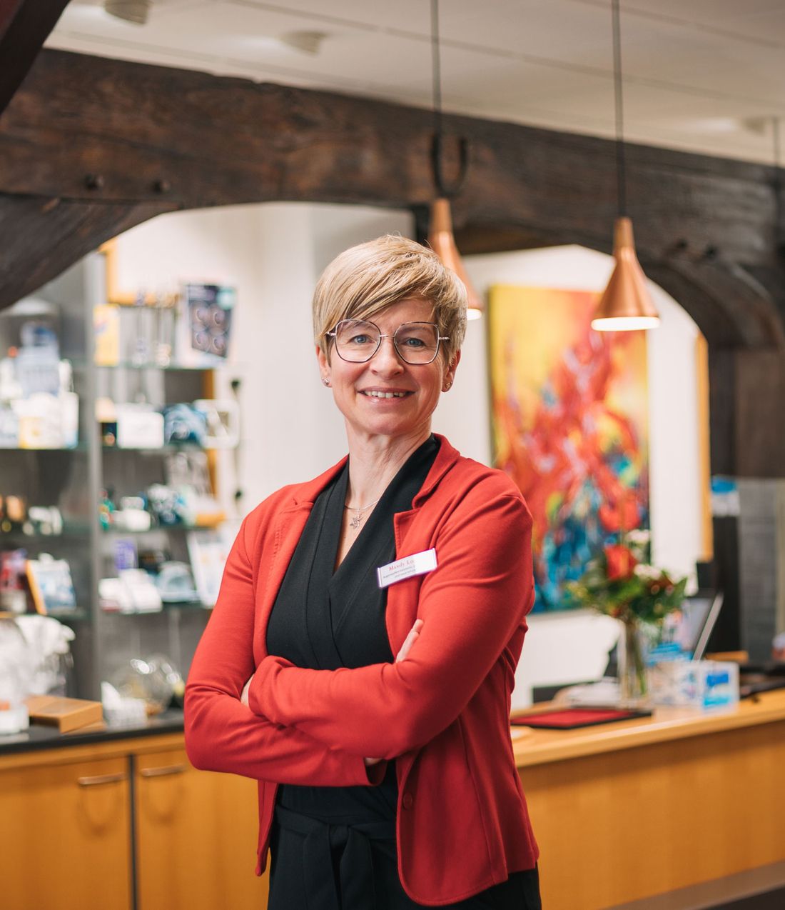 Mandy Lorenz in einer roten Strickjacke steht selbstbewusst mit verschränkten Armen in einem modernen Büro mit dekorativen Kunstwerken im Hintergrund.